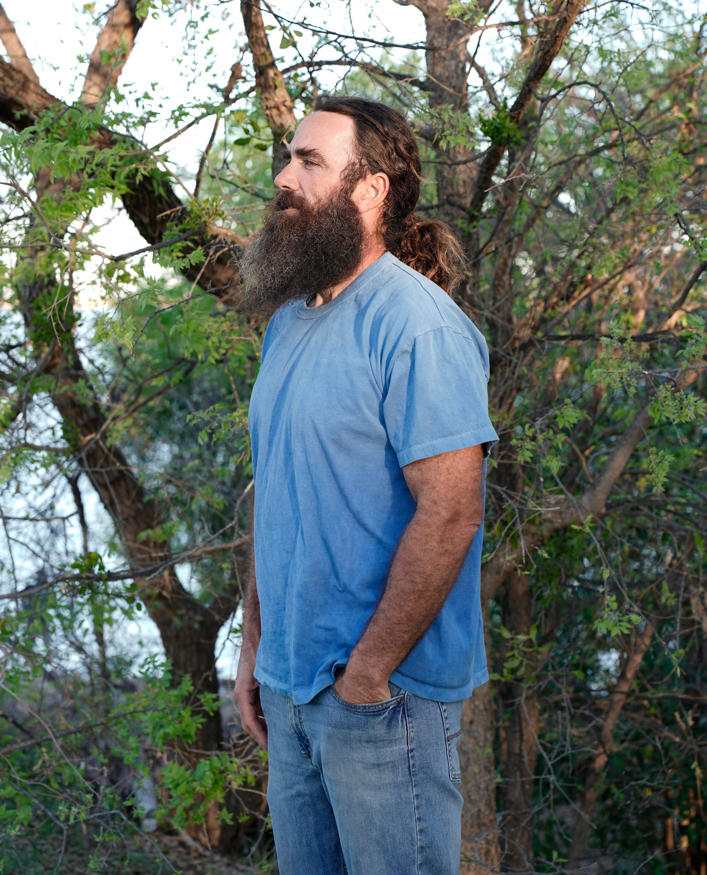 Man with a beard and long hair wearing a blue organic cotton shirt and jeans standing in a forest.