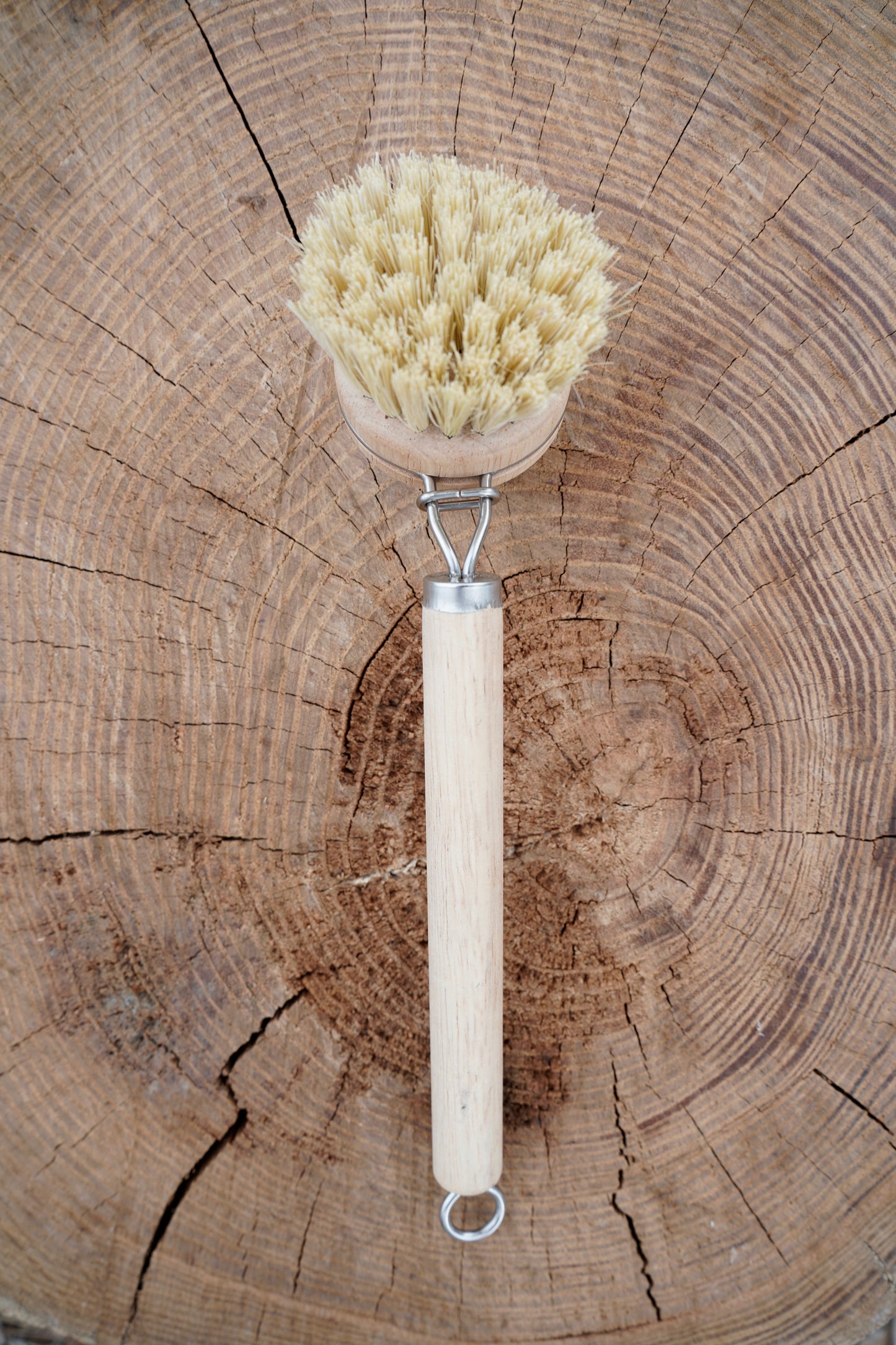 A natural sisal bristle brush with a light wood handle and metal loop, placed on a rustic wood log background.