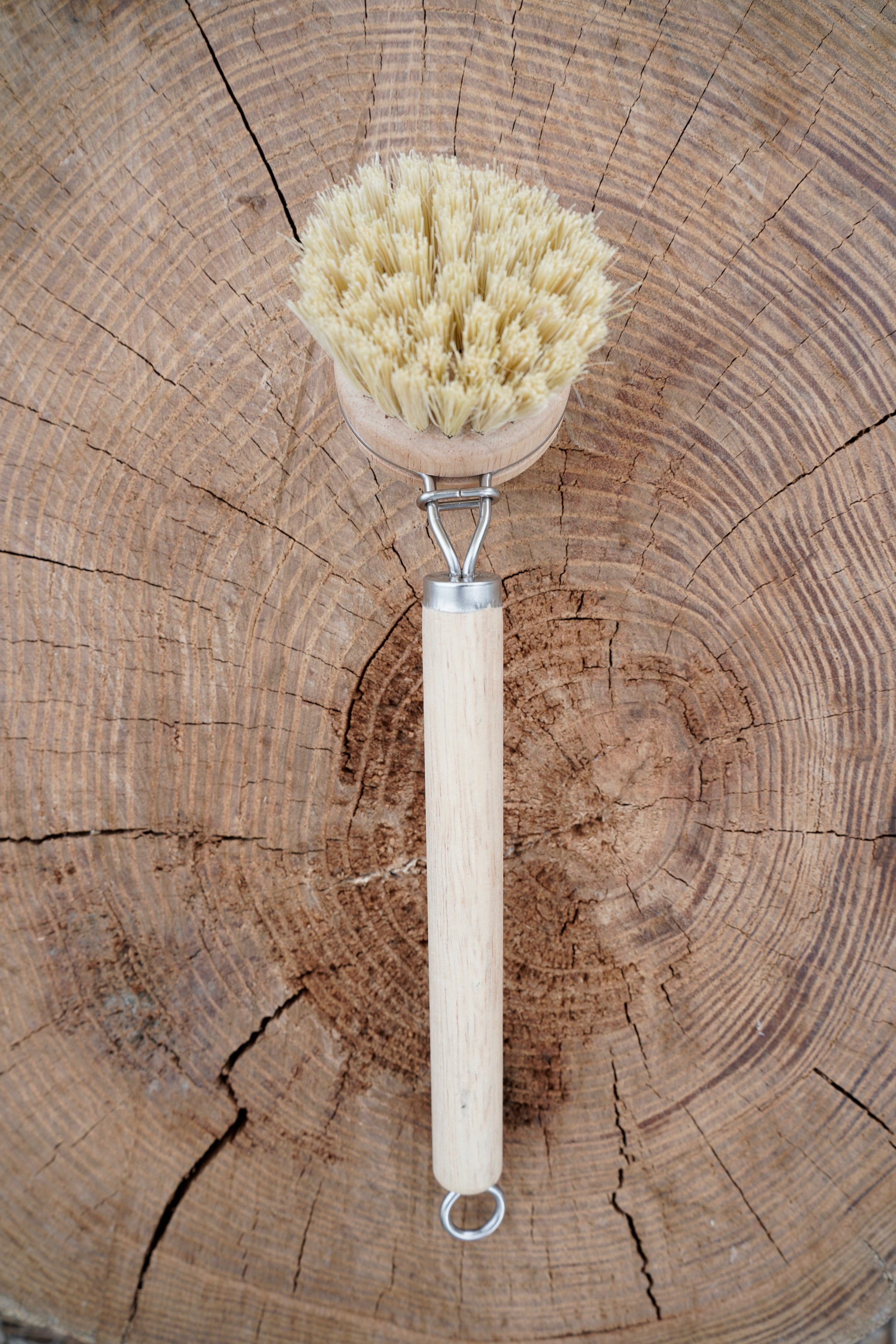 A natural sisal bristle brush with a light wood handle and metal loop, placed on a rustic wood log background.
