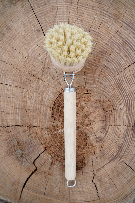 A natural sisal bristle brush with a light wood handle and metal loop, placed on a rustic wood log background.
